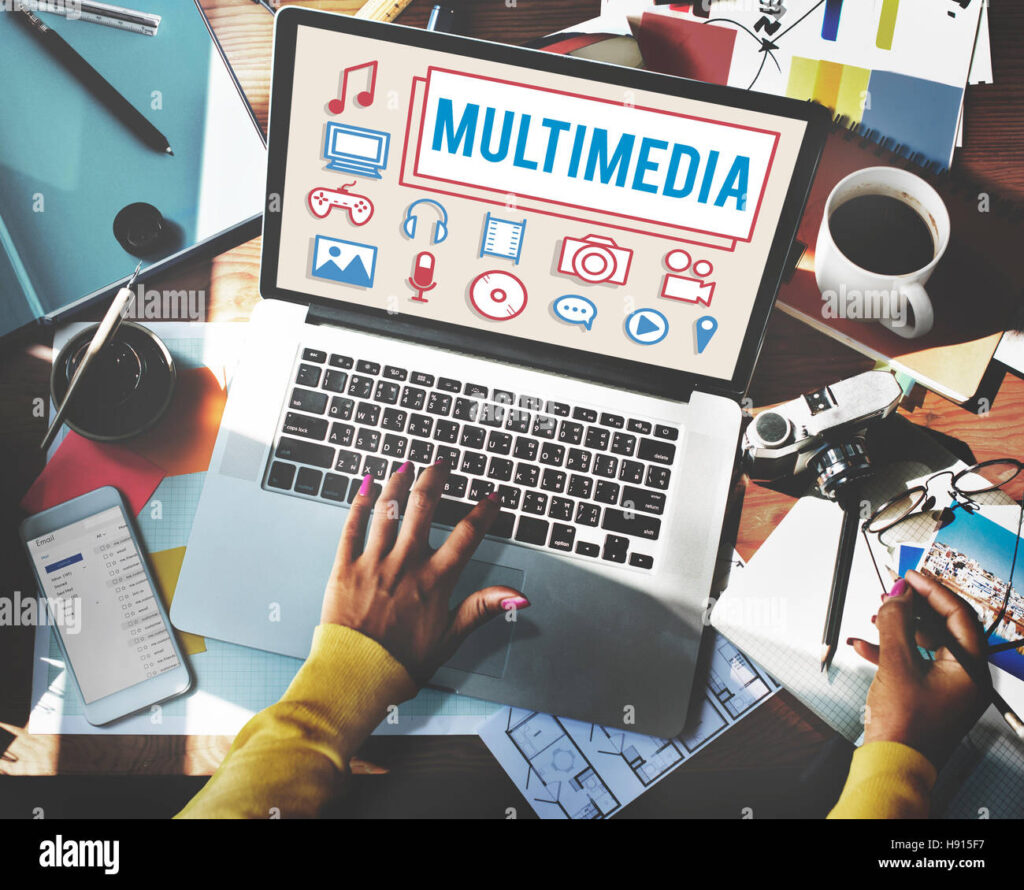 Person working on a laptop displaying the word "MULTIMEDIA" with various multimedia icons, surrounded by a smartphone, camera, coffee cup, and creative workspace items.