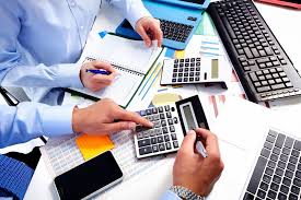 Two people in light blue dress shirts working together at a desk filled with financial tools, including two calculators, a laptop, a tablet, a notebook, and various charts. their doing accounting